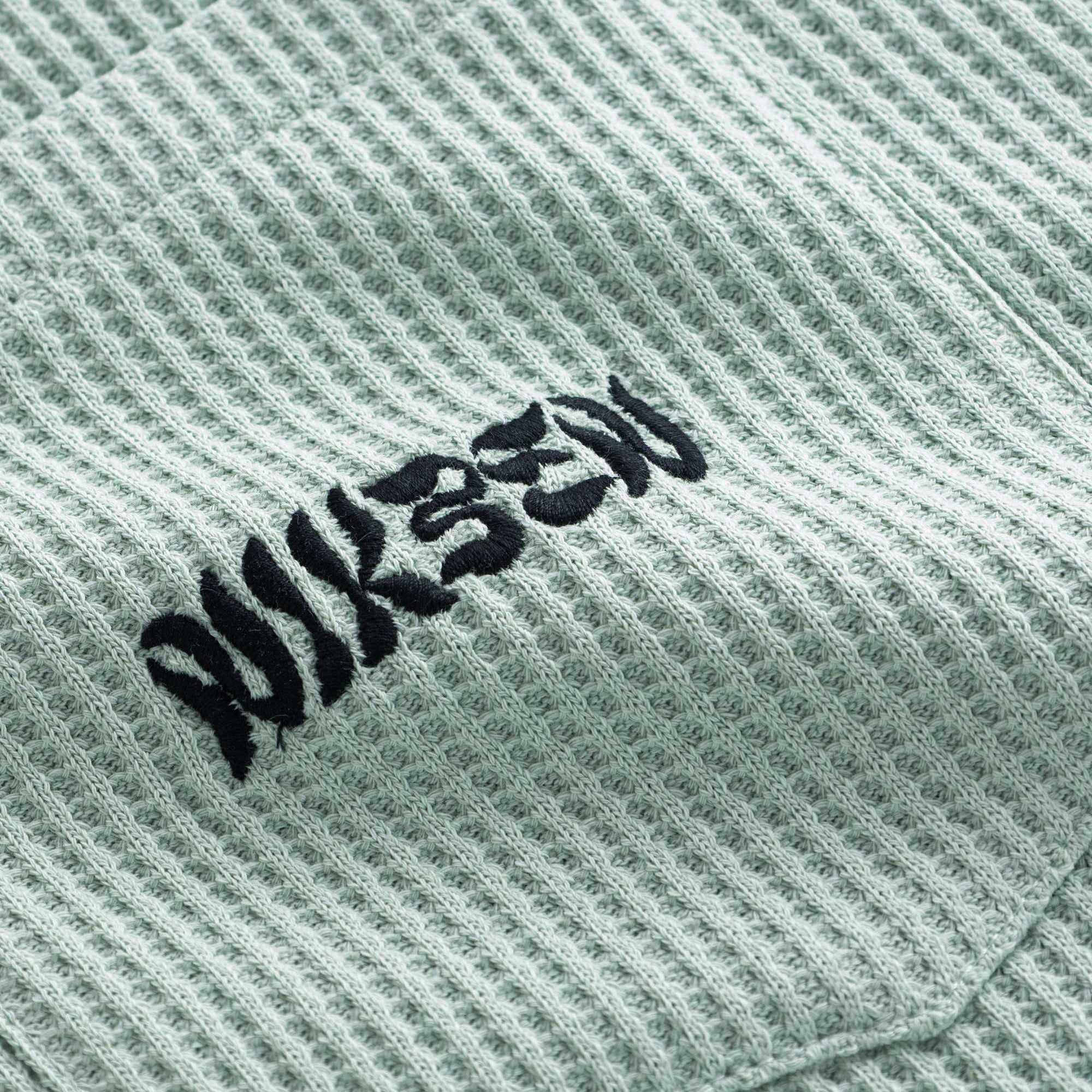 Close-up view of front pocket with stitched black logo on a mint green waffle-patterned shirt