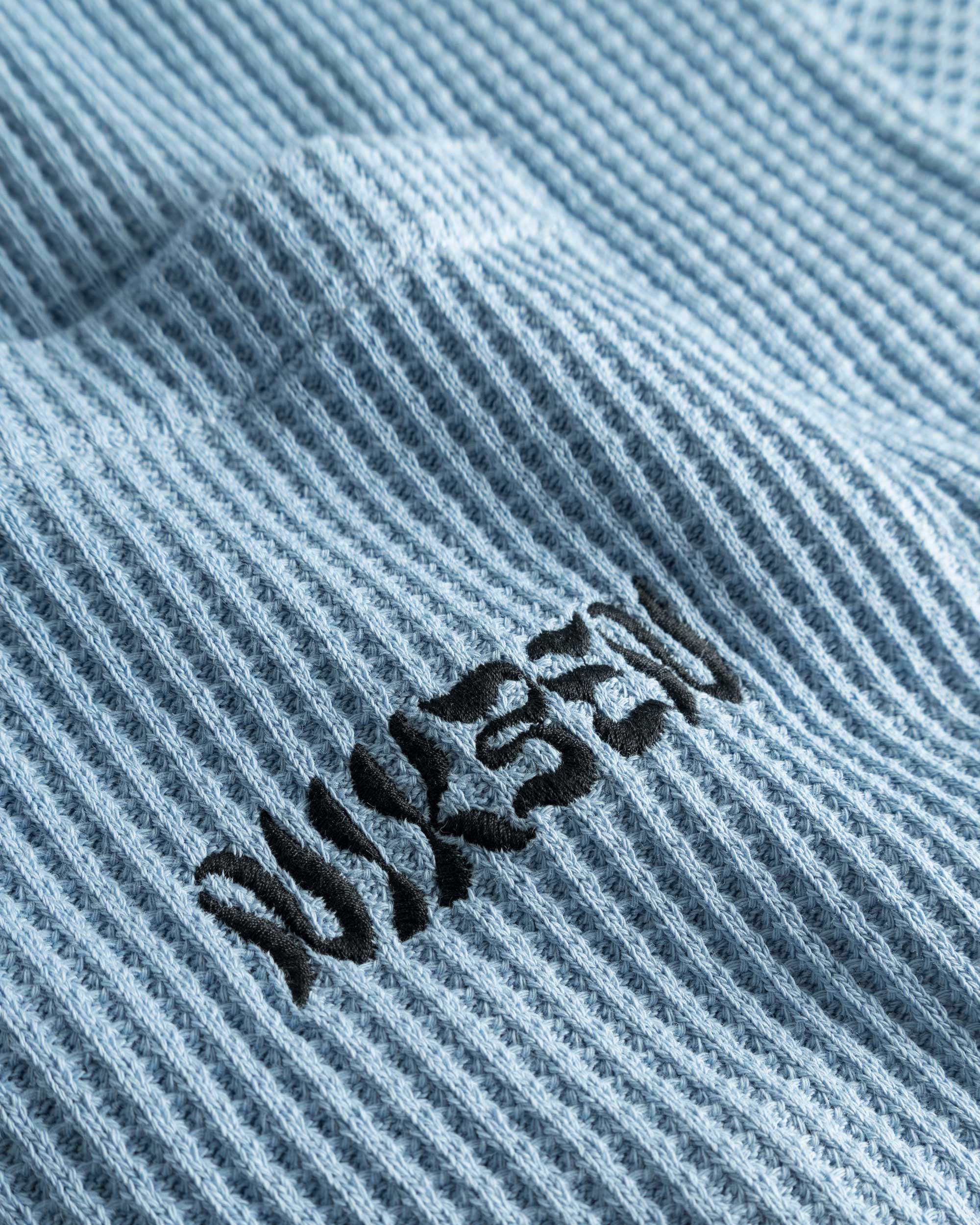 Close-up view of front pocket with stitched black logo on a sky blue waffle-patterned shirt