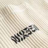 Close-up view of front pocket with stitched black logo on an off white waffle-patterned shirt