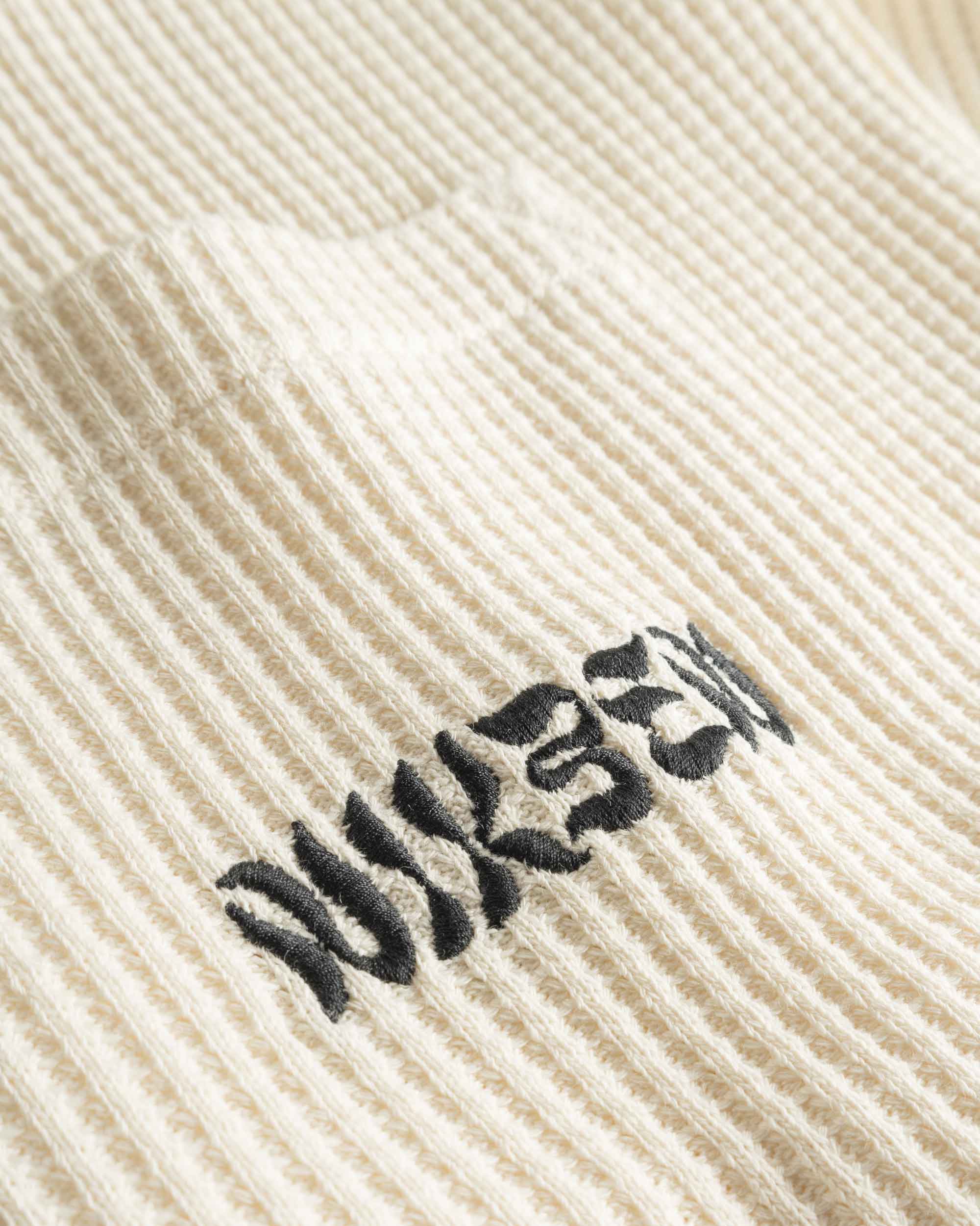 Close-up view of front pocket with stitched black logo on an off white waffle-patterned shirt