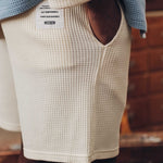 Back view of male model wearing off white waffle-patterned mid-length shorts with two front pockets and a drawstring.