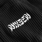 Close-up view of front pocket with stitched white logo on a black waffle-patterned shirt.