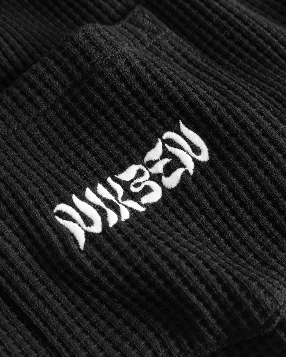 Close-up view of front pocket with stitched white logo on a black waffle-patterned shirt.